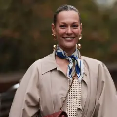 Uśmiechnięta kobieta w beżowym płaszczu i stylowej apaszce z niebieskimi wzorami. Inspiracja, jak nosić apaszkę, by dodać elegancji.