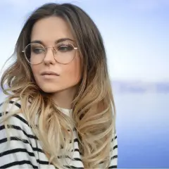 Blondynka w okularach o okrągłych oprawkach, w pasiastym swetrze, patrzy w bok na tle błękitnego nieba i wody.