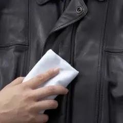 Dłoń z białą ściereczką delikatnie czyści czarną kurtkę skórzaną, pokazując, jak wyczyścić kurtkę skórzaną.