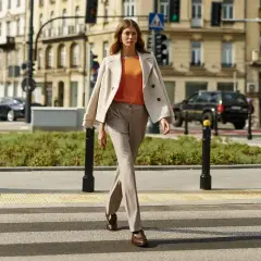 Elegancka kobieta w beżowym płaszczu i pomarańczowej bluzce przechodzi przez pasy. Stylowe stylizacje do beżowego płaszcza na co dzień.