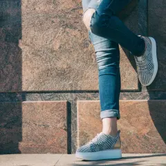 Dziewczyna w podartych jeansach i modnych sneakersach z wzorem pinroll opiera się o kamienną ścianę.