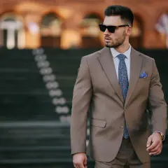 Elegancki mężczyzna w brązowej marynarce, z niebieską poszetką w kratkę i wzorzystym krawatem, w okularach przeciwsłonecznych.