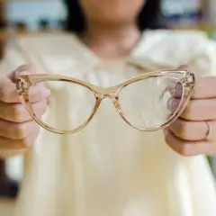 Dłonie trzymają transparentne okulary w beżowej oprawce. Podpowiedź, jak dobrać okulary, by pasowały do kształtu twarzy.
