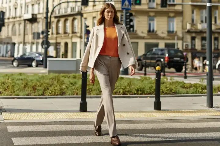 Elegancka kobieta w beżowym płaszczu i pomarańczowej bluzce przechodzi przez pasy. Stylowe stylizacje do beżowego płaszcza na co dzień.