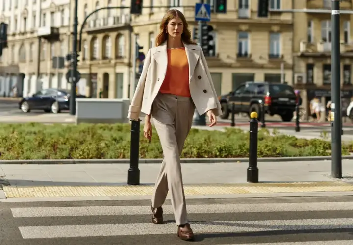 Elegancka kobieta w beżowym płaszczu i pomarańczowej bluzce przechodzi przez pasy. Stylowe stylizacje do beżowego płaszcza na co dzień.