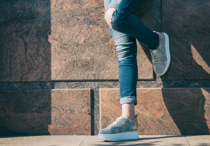 Dziewczyna w podartych jeansach i modnych sneakersach z wzorem pinroll opiera się o kamienną ścianę.