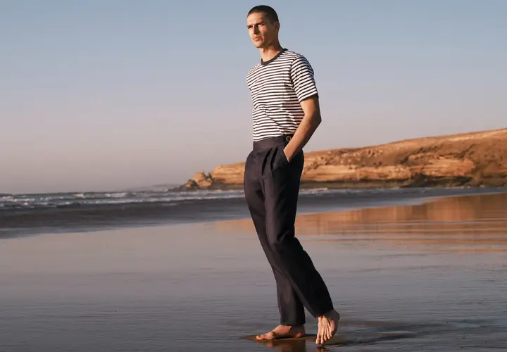 Mężczyzna w granatowych chinosach i pasiastym T-shircie spaceruje po plaży. Chinosy co to? To uniwersalne spodnie na każdą okazję.