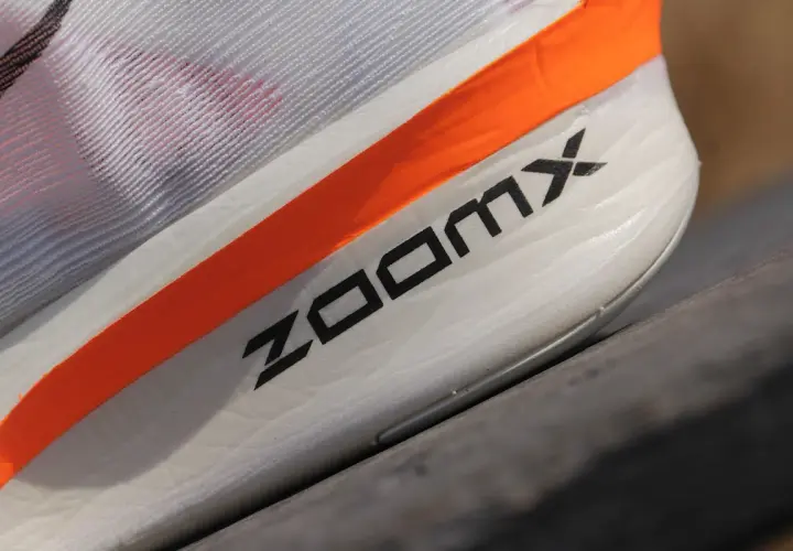 Biały but sportowy z pomarańczowym paskiem i napisem ZOOMX.
