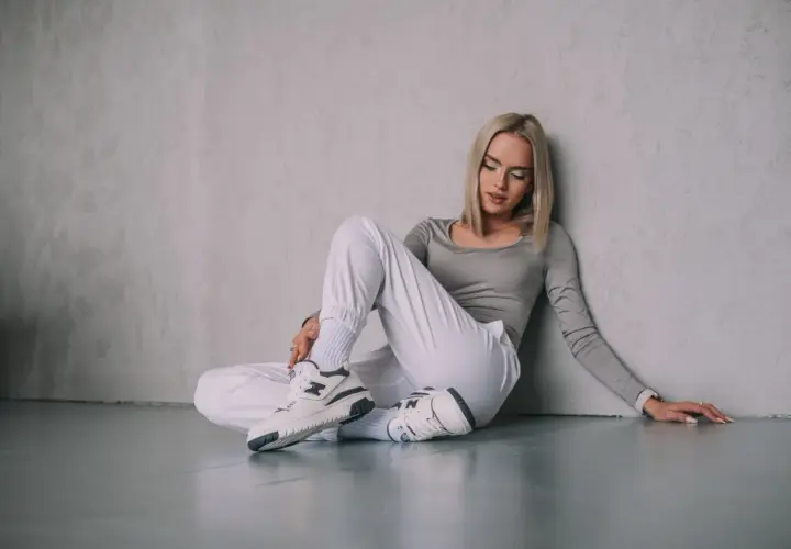Blondynka w białych spodniach i szarym topie siedzi na podłodze, opierając się o ścianę. Na nogach ma stylowe snekarsy.