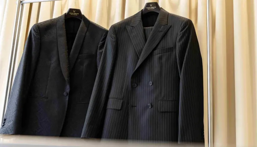Dwa eleganckie garnitury wiszą na wieszakach. Jeden w prążki, drugi z subtelnym wzorem. Prasowanie garnituru to klucz do nienagannego wyglądu.