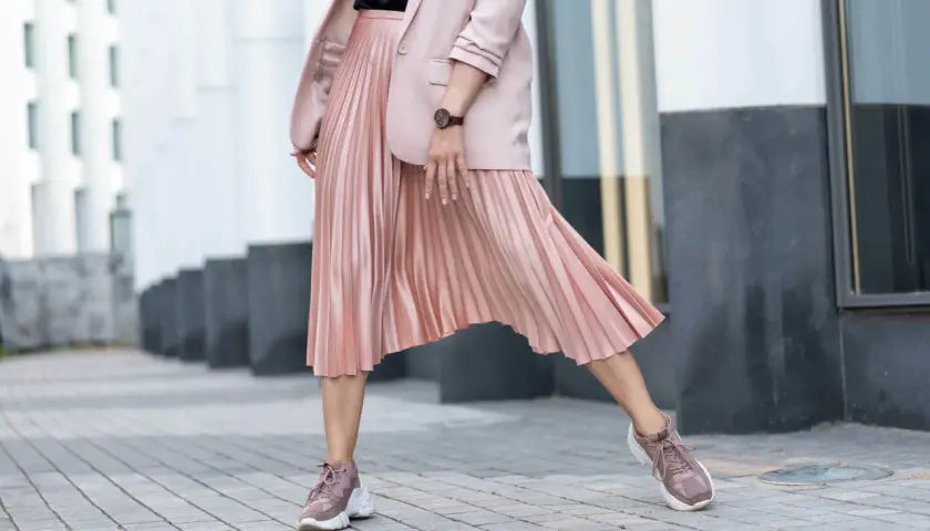 Kobieca stylizacja: pudrowy różowy żakiet i plisowana spódnica. Jakie buty do plisowanej spódnicy? Sneakersy w tym samym kolorze tworzą modny look.