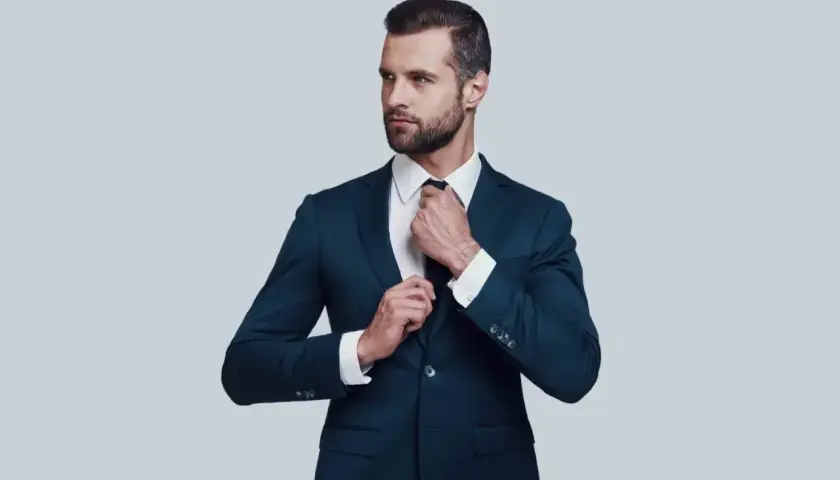 Elegancki mężczyzna w granatowym garniturze i białej koszuli poprawia krawat. Idealna stylizacja, gdy zastanawiasz się, jaka koszula do granatowego garnituru.