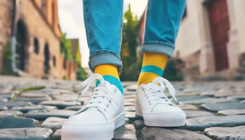 Kolorowe skarpetki do sneakersów dodają charakteru stylizacji. Osoba w jeansach i białych butach idzie po bruku.
