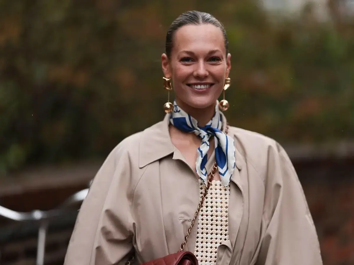 Uśmiechnięta kobieta w beżowym płaszczu i stylowej apaszce z niebieskimi wzorami. Inspiracja, jak nosić apaszkę, by dodać elegancji.