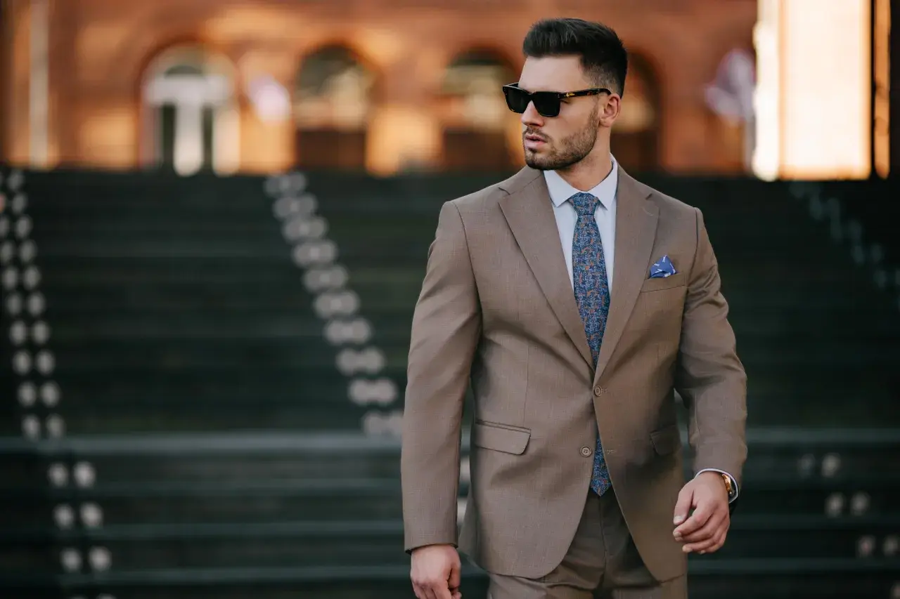 Elegancki mężczyzna w brązowej marynarce, z niebieską poszetką w kratkę i wzorzystym krawatem, w okularach przeciwsłonecznych.
