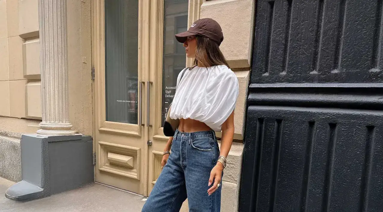 Stylowa kobieta w białej bluzce i jeansach pokazuje, jak nosić czapkę z daszkiem, tworząc modny look.