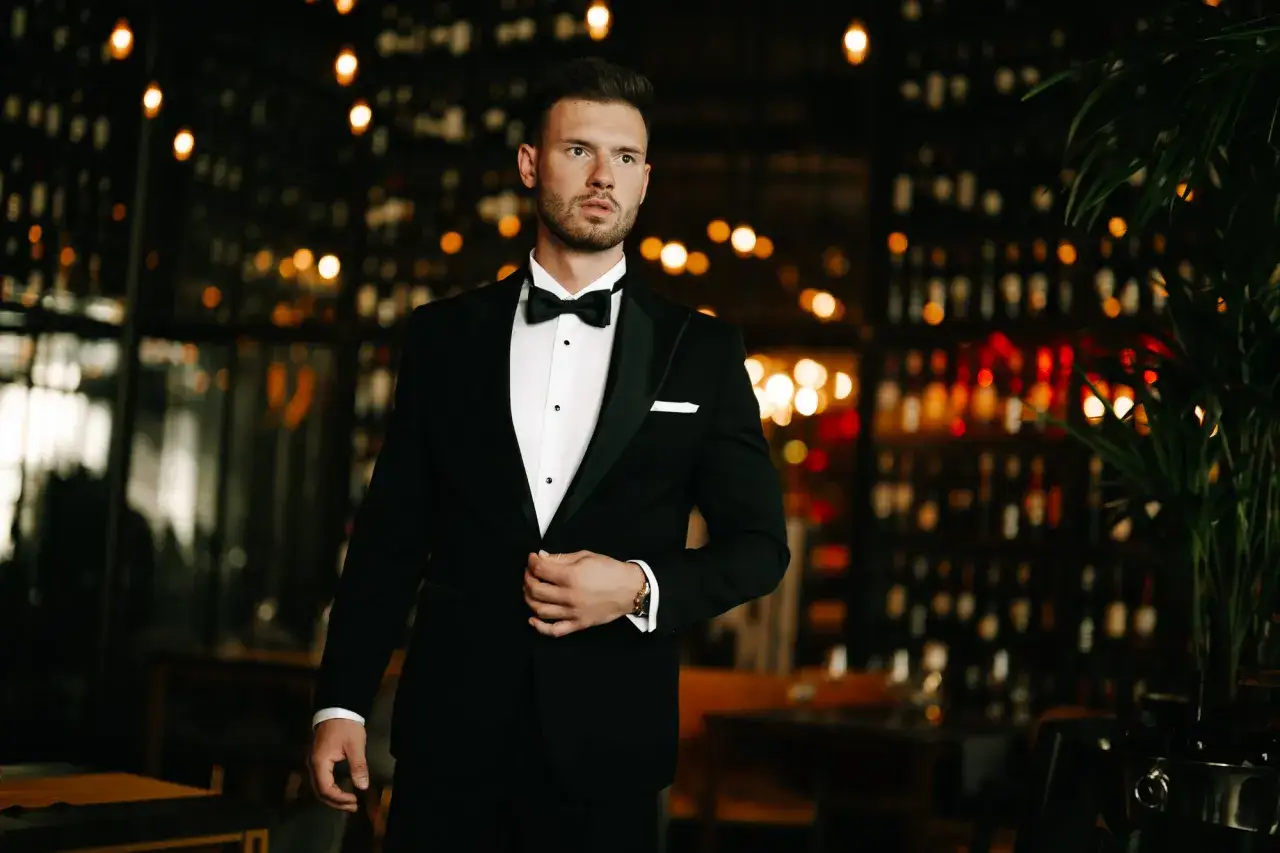 Elegancki mężczyzna w smokingu, który odróżnia się od garnituru satynowymi klapami. Tło tworzy przytulne wnętrze z butelkami wina.