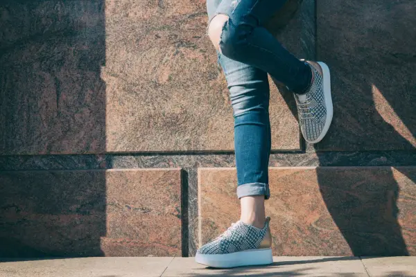 Dziewczyna w podartych jeansach i modnych sneakersach z wzorem pinroll opiera się o kamienną ścianę.